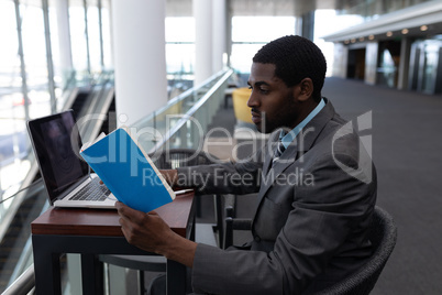 Side view of African-American businessman with laptop sitting at table and reading a book in modern