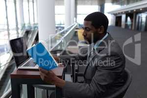 Side view of African-American businessman with laptop sitting at table and reading a book in modern