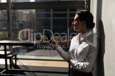 Young Caucasian businesswoman leaning against wall and using mobile phone in office