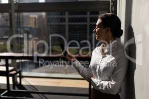 Young Caucasian businesswoman leaning against wall and using mobile phone in office
