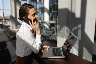 Young Caucasian businesswoman talking on mobile phone while using laptop in modern office