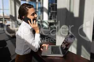 Young Caucasian businesswoman talking on mobile phone while using laptop in modern office