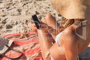 Young Caucasian woman in hat and bikini using her mobile phone on the beach