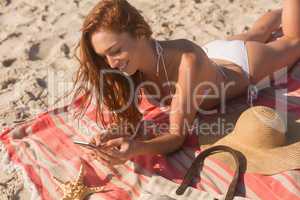 Young caucasian woman in bikini lying on the sand and using her mobile phone