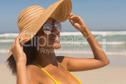 Young African American woman in yellow bikini, hat and sunglasses standing on the beach