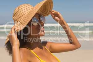 Young African American woman in yellow bikini, hat and sunglasses standing on the beach