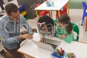 High angle view of two school girls working on one laptop while their teacher teaching them how to u