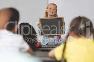 Happy Caucasian schoolgirl holding a slate in classroom at school with his classmates sitting in fro