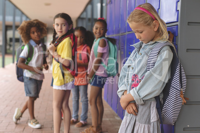 Sad schoolgirl standing in corridor while others school kids interacting with each other
