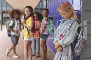 Sad schoolgirl standing in corridor while others school kids interacting with each other