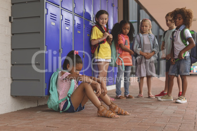 Schoolgirl sitting alone in school corridor while others school kids looking at her