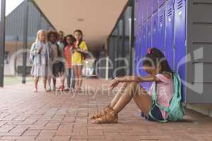 Schoolgirl sitting alone in school corridor while others school kids looking at her