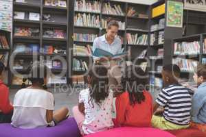 Teacher reading a story in a library to school kids sitting over big colored cushions