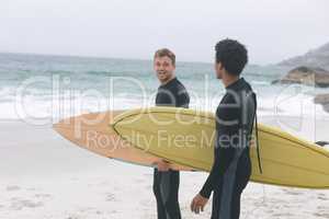 Male friends interacting with each other on beach while holding surfboard