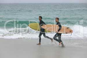 Male friends running on beach while holding surfboard