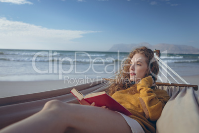 Woman holding book while lying on hammock at beach