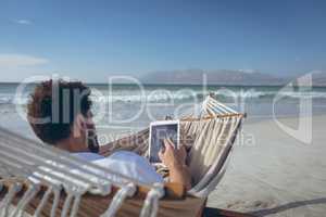Man using digital tablet while lying on hammock at beach