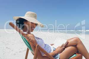Young woman holding book while sitting on sun lounger at beach