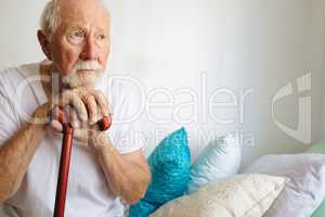 Senior male patient sitting upset on bed with stick at retirement home