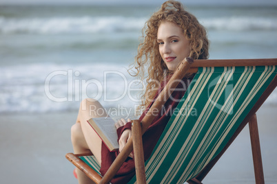 Woman holding book while sitting on sun lounger at beach