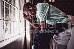 Side view of serious senior man sitting on bed by window