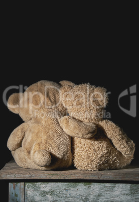 two old brown teddy bears sit hugging on a wooden surface