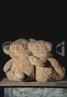 two old brown teddy bears sit hugging on a wooden surface