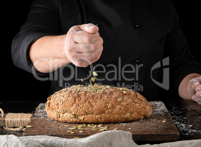 baker in black uniform sprinkles pumpkin seeds whole baked bread