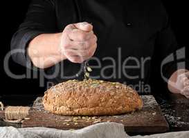 baker in black uniform sprinkles pumpkin seeds whole baked bread