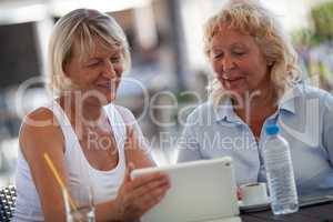 Two smiling senior women at a table of an outdoor cafe looking at the tablet screen