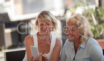 Two middle aged women sitting outside and smiling while looking at a smartphone screen