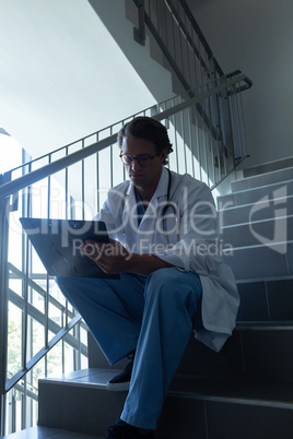 Male surgeon writing on clipboard while sitting on stairs at hospital