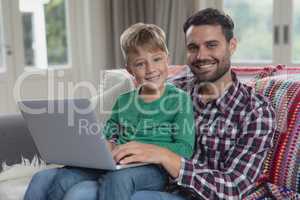 Father and son looking at camera while using laptop on sofa in a comfortable home