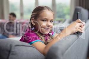 Cute girl looking at camera while using mobile phone on sofa in a comfortable home