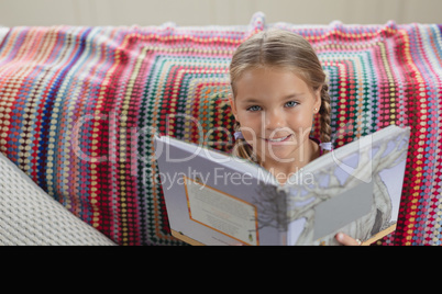 Cute girl looking at camera while reading a book on sofa in a comfortable home