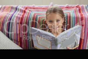 Cute girl looking at camera while reading a book on sofa in a comfortable home