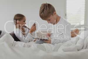 Siblings using digital tablet on bed in bedroom at comfortable home