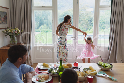 Mother dancing with her daughter while father and son sitting at dining table