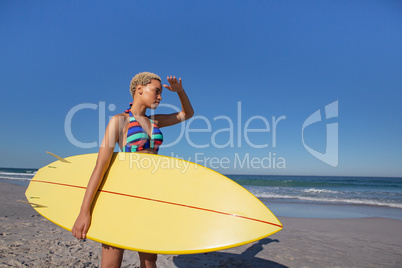 Beautiful woman in bikini with surfboard shielding eyes on beach in the sunshine