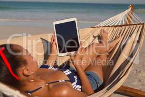 Woman using digital tablet while relaxing on hammock on beach