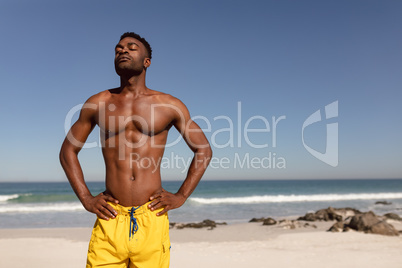 Shirtless man with hands on hip and eyes closed standing on beach in the sunshine