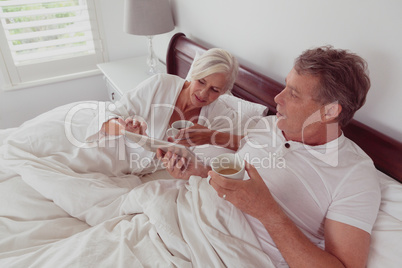 Active senior couple using digital tablet while having coffee together in bed in bedroom