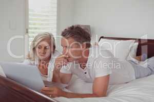 Active senior couple using laptop while lying on bed in bedroom at comfortable home