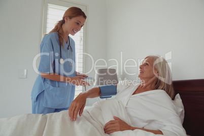 Female doctor checking blood pressure of active senior woman in bed in bedroom