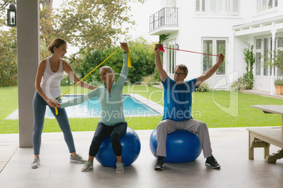 Female trainer assisting active senior couple to exercise with resistance band in the porch
