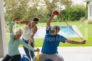 Female trainer assisting active senior couple to exercise with resistance band in the porch