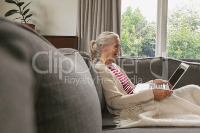 Active senior woman relaxing on sofa and using laptop in living room at comfortable home