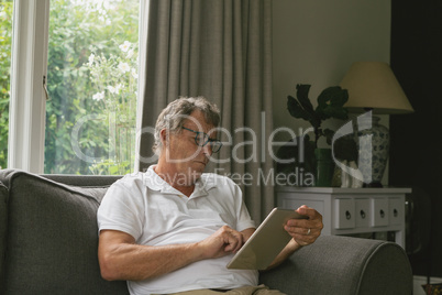 Active senior man sitting on sofa and using digital tablet in living room at comfortable home