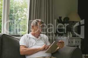 Active senior man sitting on sofa and using digital tablet in living room at comfortable home