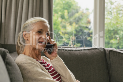 Active senior woman sitting on sofa and talking on mobile phone in living room at comfortable home
